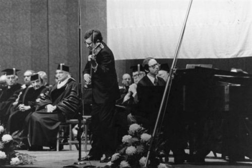 Zvi Zeitlin performs on violin with Barry Snyder at the piano during a convocation ceremony in Eastman Theatre, with honoree Golda Meir and faculty wearing regalia in the background.