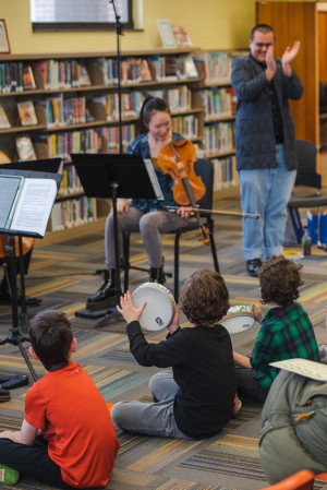 Three children sit on the carpet watching a violinist play. One child clapping a tambourine. 