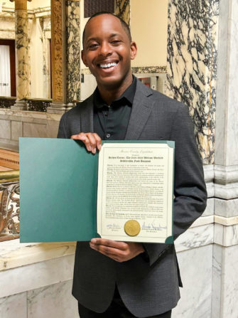 Holden Turner, an African American baritone, was honored with a Proclamation from Monroe County as the recipient of the 2024-25 William Warfield Scholarship Fund recipient. He is pictured at the Monroe County Legislative Camber, smiling in a gray suit, holding the proclamation document.