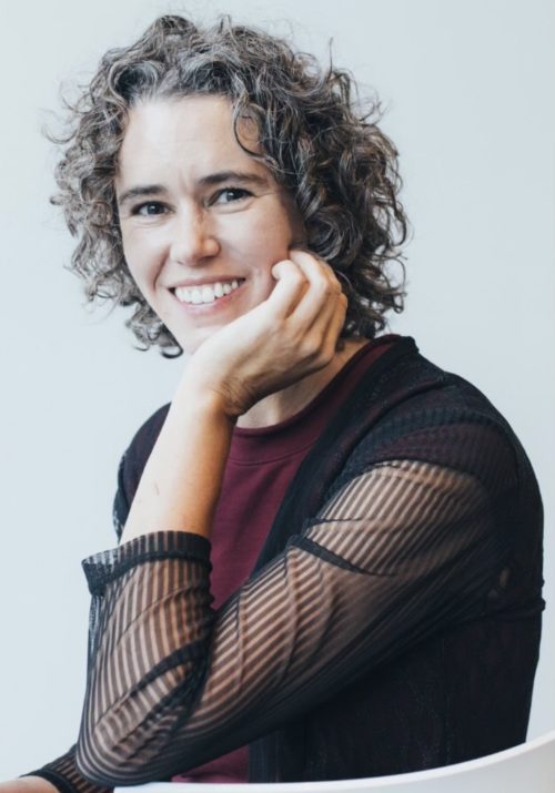 Professional headshot of conductor Matilda Hofman in a black blouse over red shirt, staring and smiling into camera. 