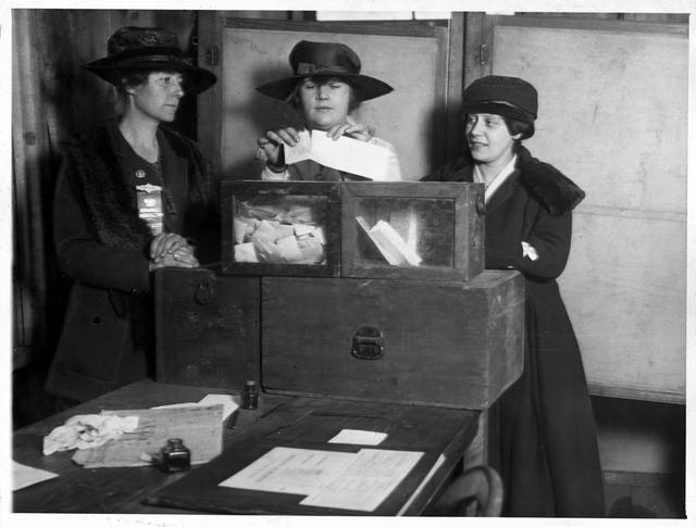 Three suffragettes standing around a tabletop ballot box casting their votes